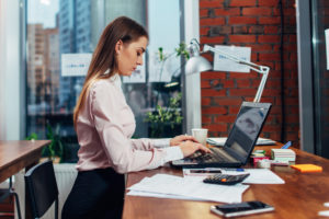 White woman with brown hair typing on a black laptop at a brown desk