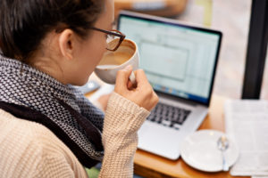 Woman with black hair wearing a black and white scarf and beige sweater sips coffee while watching an upload box on her laptop screen