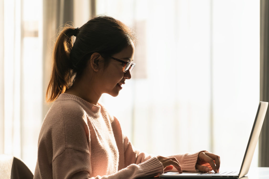 Girl with ponytail, glasses, and cozy pink sweater types on silver laptop