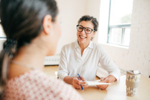 a middle-aged white woman in glasses and a white collared shirt holds a paycheck in her hand and smiles across the table at a dark-haired woman