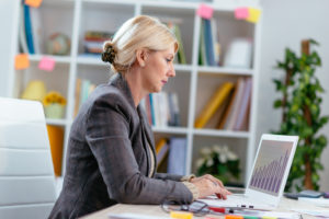 A white woman with blonde hair wearing a grey jacket sits at a desk typing on a white laptop