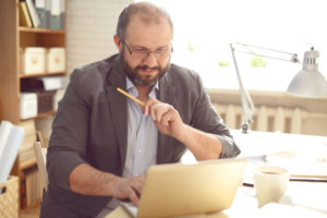 A middle-aged white man with glasses in a grey suit coat types on a white laptop while holding a pencil