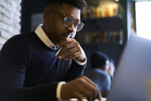 A Black man wearing glasses and a navy sweater over a white collared shirt sits in front of a laptop