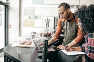Two people stand at a coffeeshop countertop, one looking at a laptop and the other doing calculations on a piece of paper