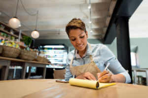 A woman with short brown hair wearing an apron writes on a yellow pad of paper at a restaurant