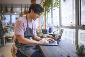 Portrait of young businessman using laptop at table. Smiling male entrepreneur is in store. He is wearing apron.