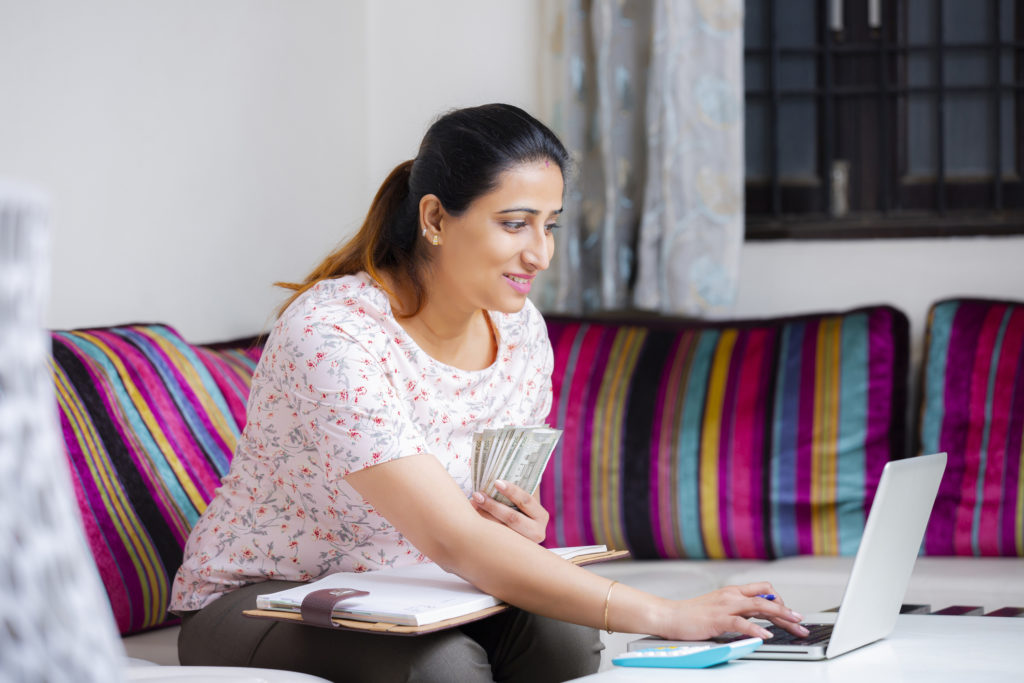 A young woman of color sits on a couch and types into a laptop while holding cash