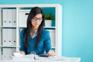A woman with long black hair and glasses flips through financial records with a calculator