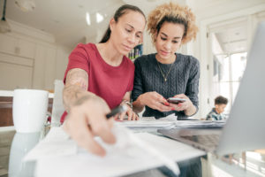 Two women look at a computer and various financial documents while crunching numbers on a calculator