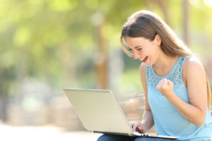 Featured image of a white woman in a blue tank top pumping her fist and cheering as she reads good news on her laptop