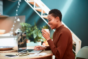 Feature image of a young woman of color sitting in front of a laptop, smiling and drinking from a mug