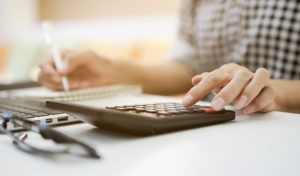 Feature image of a white woman typing on a calculator with one hand and writing on a notepad with the other