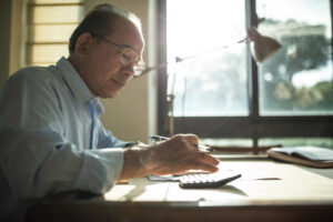 Featured image of an older Asian man sitting at a desk typing on a calculator