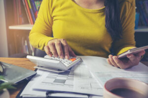 Featured image of a young woman in a yellow shirt typing on a calculator while looking at financial papers
