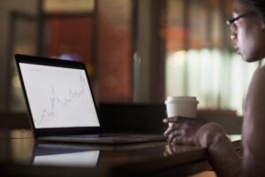 Feature image of a young Black woman drinking a coffee while looking at financial reports on her laptop