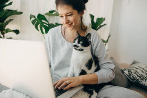 Featured image of a white woman sitting on a couch, typing on her laptop and holding a black and white cat