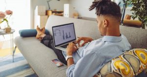 Young Black man in blue shirt reclines on white couch, looking at charts on a laptop