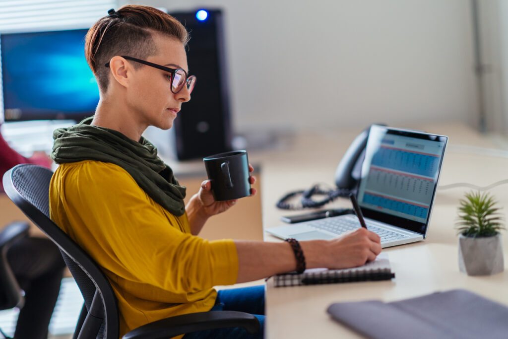 Image of a woman writing notes on a pad of paper while looking at a laptop