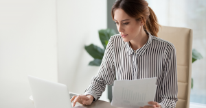 A white woman with brown hair sits at a desk, typing on a white laptop