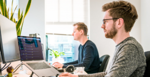 Two white men with red hair sit at separate desks typing on separate laptops