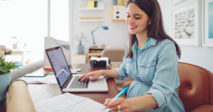 A pregnant white woman with brown hair types on a laptop with one hand and writes with a blue pencil with the other