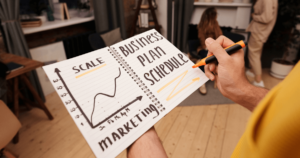 A person holds a notebook with a graph noting scale with "Marketing" on the x-axis with "Business Plan Schedule" written on the opposite page.