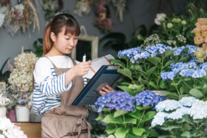 A young Asian woman in a white shirt and brown apron looks at an inventory clipboard while evaluating plants at her flower shop