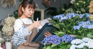 A young Asian woman looks at a list of her inventory while looking at blue flowers in her flower shop