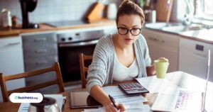 woman wearing glasses and working at kitchen table using calculator